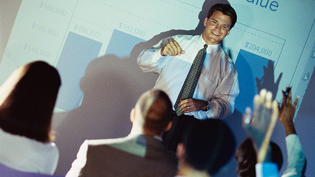 A man giving a presentation to coworkers in front of a projection screen and the audience is raising their hands asking questions.