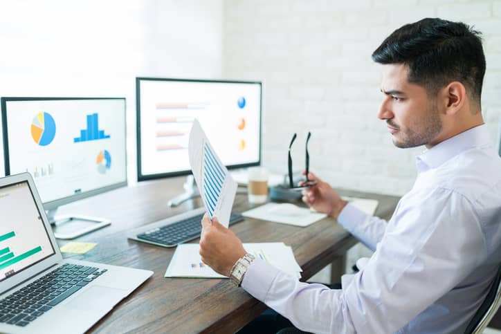 Business analyst analyzing business performance data while holding paper at desk
